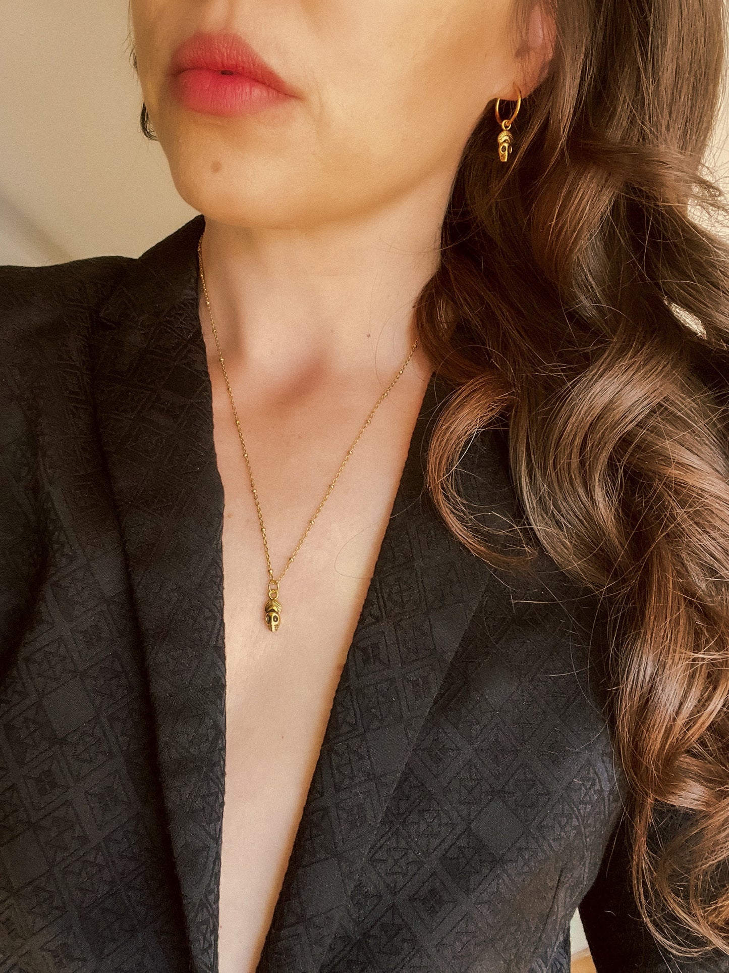 Close-up of a person wearing gold skull earrings and a skull necklace with a neutral background