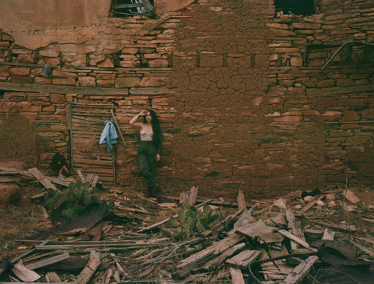 Leah Prada standing against the weathered adobe and stone wall of a ruined New Mexico structure, surrounded by fallen wood and desert vegetation.