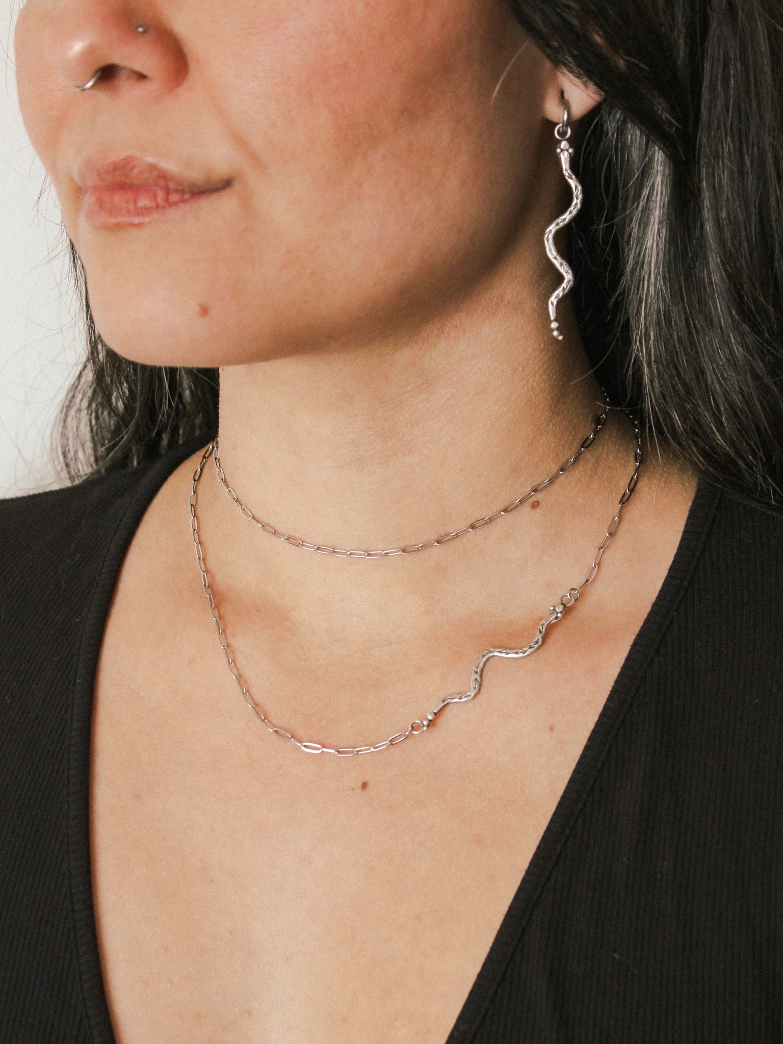 Close-up of a person wearing silver snake necklaces and earrings against a neutral background
